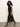 A woman with curly hair wears the Sanctuary Clothing everyday satin dress in black olive, paired with tan suede ankle boots. She walks left against a plain light background, appearing relaxed and stylish.