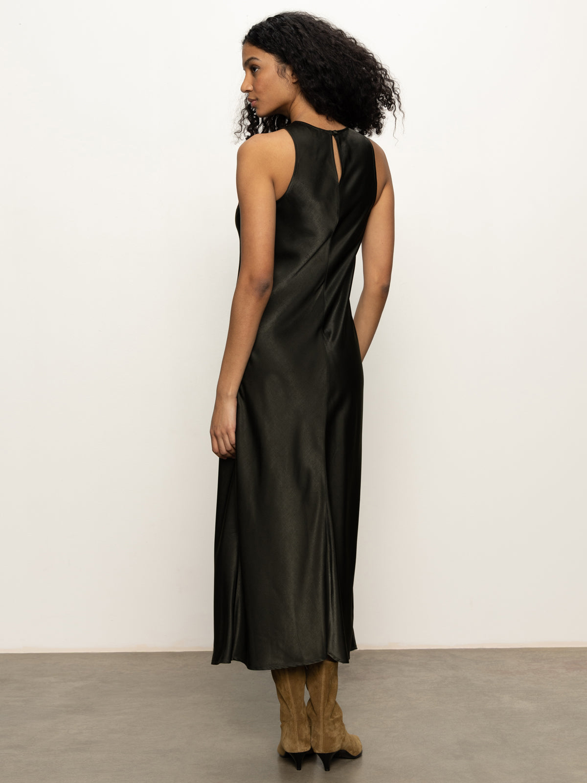 A woman with curly hair, seen from the back, wears Sanctuary Clothing's everyday satin dress in black olive and tan suede knee-high boots, standing against a plain white background.