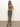 A woman poses against a white wall, wearing Sanctuary Clothing’s effortless slip dress meow—an ankle-length, sleeveless leopard print dress—with brown platform sandals. She has long brown hair and rests one hand on her other wrist.