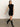 A woman walks indoors against a plain light wall and concrete floor, wearing Sanctuary Clothing's "an affair to remember dress black" with cap sleeves and knee-high black boots.