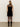 A woman with straight blonde hair wears the Sanctuary Clothing party shift dress in black, featuring a sleeveless design with sheer lace at the upper back. She pairs it with black sheer tights and knee-high boots, standing with her back to the camera.