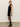 A woman in profile wears the Sanctuary Clothing party shift dress black with a high neckline and sheer details, paired with black tights, ankle boots, sunglasses, and straight hair down against a plain background.