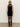 A woman wearing the Sanctuary Clothing party shift dress in black stands against a plain light wall, styled with black tights, knee-high boots, and dark sunglasses.
