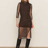 A woman poses against a plain light background in the Sanctuary Clothing night out mock sweater dress semi sweet, a fitted black dress with three-quarter sleeves and side slit, paired with knee-high black boots.