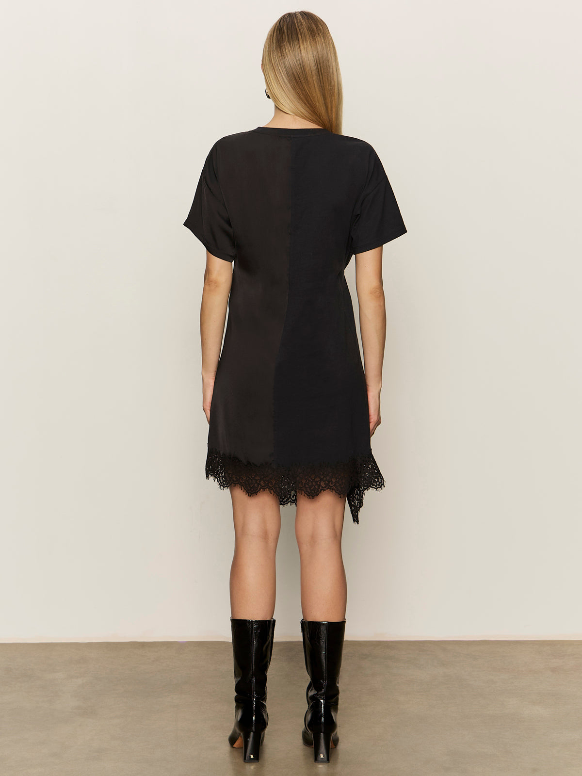 A woman with long blonde hair faces away, wearing the Sanctuary Clothing satin mix t-shirt dress in black, paired with black knee-high boots, against a plain light background.
