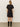 A woman with long blonde hair faces away, wearing the Sanctuary Clothing satin mix t-shirt dress in black, paired with black knee-high boots, against a plain light background.