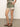 The person is wearing Sanctuary Clothing's Spring Breaker Short in Mezzo Sage, featuring patterned green fabric with pink and white designs, paired with a matching top and brown leather sandals. They stand on a concrete floor with hands in pockets, facing slightly sideways.