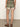 A person is seen from the rear wearing Sanctuary Clothing's Spring Breaker Short in Mezzo Sage, featuring floral patterns with a white trim, paired with tan strappy sandals. The backdrop is a plain, neutral wall and floor.