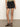 A person stands against a plain background wearing Sanctuary Clothing's view cargo short in black, featuring front pockets and button details. Paired with a green top and black ankle-strap sandals, the shorts offer a tailored fit.