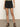 A person is standing on a gray floor wearing Sanctuary Clothing's view cargo short in black, featuring flap pockets, paired with a green top and black sandals. The backdrop is a plain, light-colored wall.