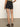 Rear view of a person wearing Sanctuary Clothing's black "view cargo short," a green top, and black sandals. The person stands on a plain surface against a light background with their right hand on their hip.