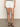 A person is wearing the Sanctuary Clothing "view cargo short" in brilliant white, featuring gold button details and front pockets. They paired it with a black and white top and brown strappy sandals, set against a plain backdrop with a concrete-colored floor.