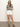 A person stands against a plain backdrop, wearing a black and white striped shirt, Sanctuary Clothing's view cargo short in brilliant white with gold buttons, and brown sandals. They have long hair and a relaxed posture with a neutral facial expression.