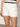 The person is wearing Sanctuary Clothing's "View Cargo Short" in brilliant white, featuring front pockets and gold buttons, paired with a black and white striped knitted top against a plain, light-colored wall.