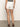 Rear view of a person wearing a black and white top, Sanctuary Clothing's "view cargo short" in brilliant white, and brown sandals on a plain surface with a neutral background. The person faces away from the camera.