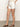 A person sports a sleeveless, knitted beige top and Sanctuary Clothing's arden denim shorts in brilliant white, with one hand on their hip. They also wear brown flat sandals, set against a plain, neutral background.