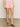 A person stands indoors against a plain wall and floor, wearing arden denim shorts in washed cherry blossom from Sanctuary Clothing, paired with a white top and brown sandals featuring gold accents. The focus is on their lower half.