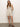 A woman in Sanctuary Clothing's caravan cuffed short flax, part of a sleeveless, buttoned tan ensemble, stands against a plain light background. Wearing brown sandals and with long brown hair, she smiles slightly while hiding her hands behind her back.