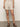A person wearing Sanctuary Clothing's Caravan Cuffed Short in Flax and brown sandals stands against a plain wall, arms relaxed by their sides. The focus is on the shorts and sandals.
