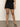 A person wearing Sanctuary Clothing's voyage short black and black heeled sandals stands on a gray floor against an off-white wall, with only their lower half visible.