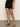 A person wearing the "Voyage Short Black" from Sanctuary Clothing, paired with a black sleeveless top and black heeled sandals, stands on a brown floor against a plain white wall, hands resting by their sides without their face visible.