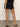 A person in a black top and Sanctuary Clothing's voyage short in black is standing with their back to the camera, wearing black heeled sandals on a concrete floor against a plain, light-colored background.