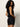 A woman with curly hair models Sanctuary Clothing's black voyage shorts and a sleeveless black top, glancing over her shoulder against a neutral background.