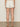 A person stands with their back to the camera, wearing Sanctuary Clothing's voyage short in bleached white, a cream knit top, and open-back sandals against a neutral floor and plain beige wall backdrop.