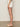 Only the lower half of a woman is visible as she stands sideways against a plain background, wearing Sanctuary Clothing’s voyage short in bleached white, paired with a white top and cream slide sandals with a low wooden heel.