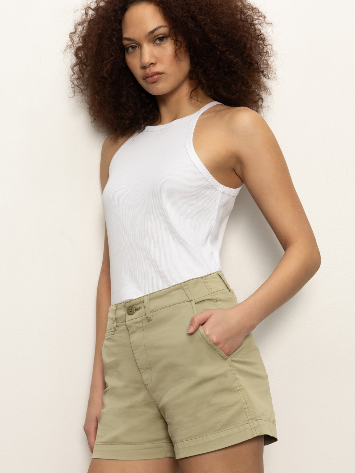 A person with curly hair wears the Sanctuary Clothing liner short bamboo, a white sleeveless top, and stands against a plain light wall with one hand in their pocket and a neutral expression.