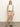 A woman with straight blonde hair and a neutral expression stands against a plain background wearing Sanctuary Clothing„s liner short in sandstone, styled with a white sleeveless top, black belt, and black sandals; one hand rests in her pocket.