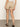 A woman stands indoors facing away from the camera, wearing Sanctuary Clothing's liner short in sandstone, a white top, and black sandals on a gray floor with a plain light-colored wall.