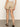 A woman stands indoors facing away from the camera, wearing Sanctuary Clothing's liner short in sandstone, a white top, and black sandals on a gray floor with a plain light-colored wall.