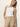 A woman with straight blonde hair wears the Sanctuary Clothing liner short in sandstone and a white sleeveless top, standing against an off-white wall with one hand in her pocket, looking into the camera.