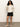 A woman with curly hair stands against a plain background, confidently looking at the camera. She wears a white zip-up knit sweater, beige slip-on shoes, and Sanctuary Clothing’s cruiser short in black, with her hands in her pockets.