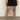 Only the lower half of a person is shown, wearing Sanctuary Clothing’s cruiser short black with a white knit top and cream woven flats, standing against a plain wall.