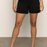Only the lower half of a person is shown, wearing Sanctuary Clothing’s cruiser short black with a white knit top and cream woven flats, standing against a plain wall.