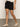 A person stands indoors with their arms relaxed at their sides, wearing Sanctuary Clothing's cruiser short in black, paired with a white textured top and cream-colored woven flats against a plain, neutral background.