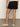A person faces away indoors, wearing Sanctuary Clothing cruiser short black with back pockets, a cream knit top, and beige woven slip-ons. The background is plain, featuring a gray floor and light-colored wall.