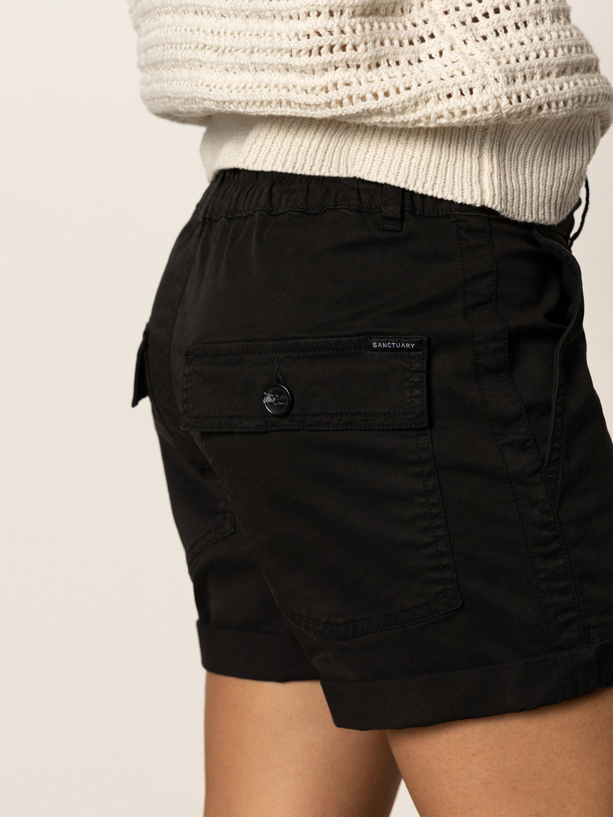 A person shown from the waist down and side, wearing Sanctuary Clothing's cruiser short black with a buttoned back pocket and a white knitted sweater.