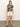 A woman stands against a plain background, wearing a black and beige abstract print t-shirt, the Sanctuary Clothing cruiser short in washed sandstone, and brown sandals. She has her hands in her pockets and looks directly at the camera.