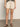 A person stands on a smooth floor against a plain light wall, wearing Sanctuary Clothing's cruiser short washed sandstone, a black and white patterned top, and brown sandals.