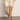 A person stands on a smooth floor with hands in pockets and legs crossed at the ankle, wearing Sanctuary Clothing’s cruiser short in washed sandstone. Their upper face is not visible.