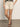 A person stands on a smooth floor with hands in pockets and legs crossed at the ankle, wearing Sanctuary Clothing’s cruiser short in washed sandstone. Their upper face is not visible.