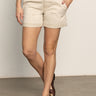 A person stands on a smooth floor with hands in pockets and legs crossed at the ankle, wearing Sanctuary Clothing’s cruiser short in washed sandstone. Their upper face is not visible.