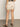 A person is indoors wearing the Sanctuary Clothing cruiser short in washed sandstone, featuring back pockets with black buttons. Brown sandals and a patterned top are also visible in this rear, waist-down view.