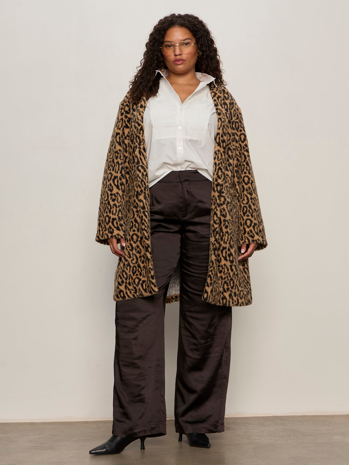 A woman with curly hair wears glasses, a white button-up, loose black pants, black heeled boots, and the Sanctuary Clothing hometown jacket lynx extended sizing, standing against a plain light wall facing forward.