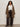 A woman with curly hair wears glasses, a white button-up, loose black pants, black heeled boots, and the Sanctuary Clothing hometown jacket lynx extended sizing, standing against a plain light wall facing forward.