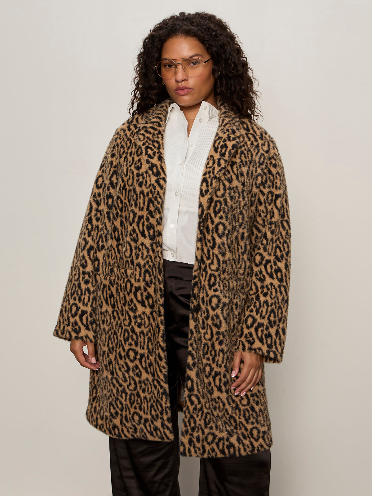 A person with curly hair wears glasses, a white button-up shirt, dark pants, and Sanctuary Clothing„¢s hometown jacket lynx extended sizing while standing against a plain light background.
