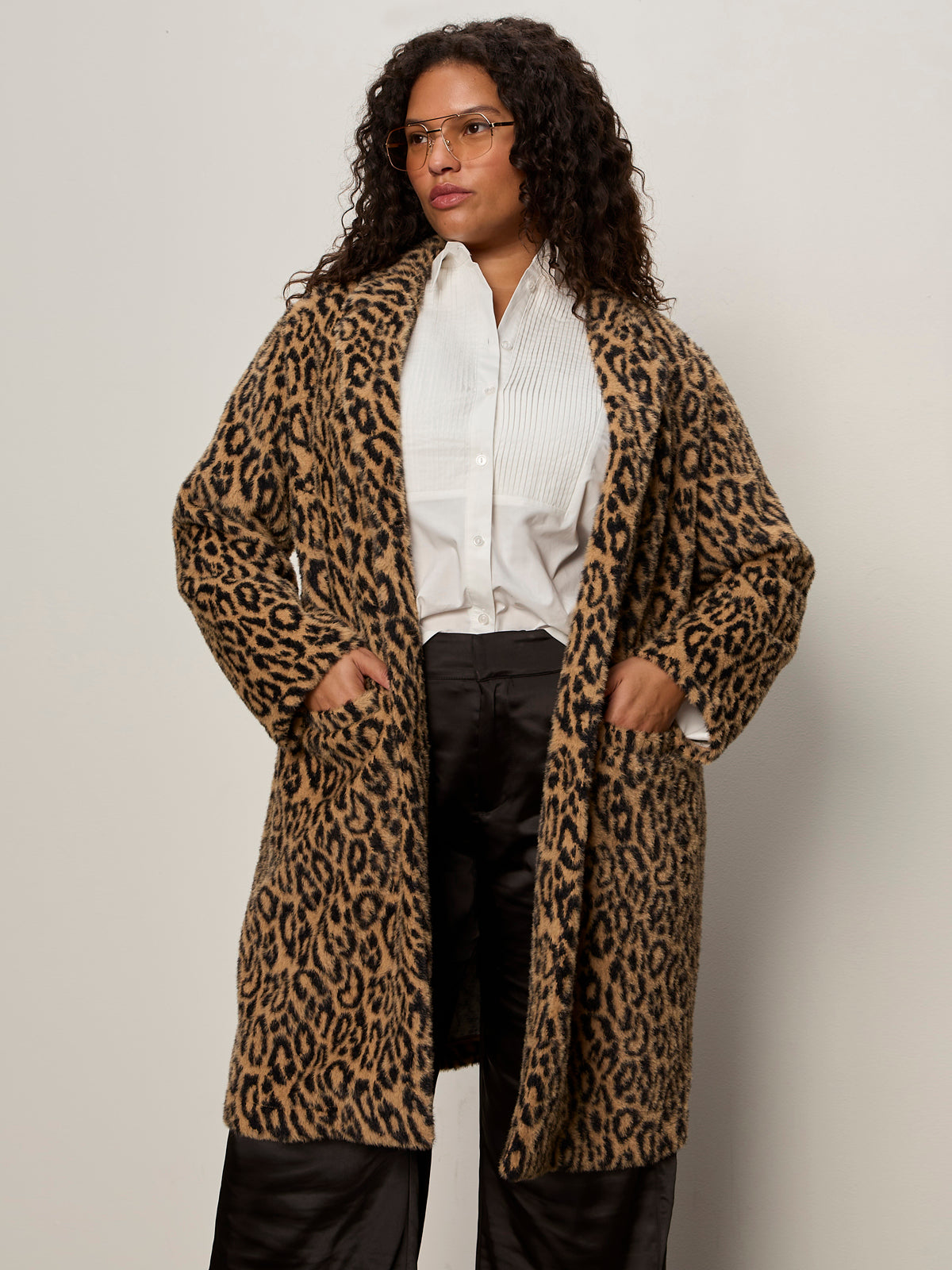 A woman with curly hair and glasses stands against a plain background, wearing the Sanctuary Clothing hometown jacket lynx extended sizing over a white button-up shirt, black satin pants, and keeps her hands in the coat pockets.